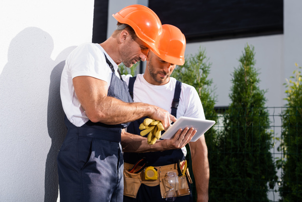 Handyman in overalls using digital tablet while standing near facade of house  - Foto, Imagen