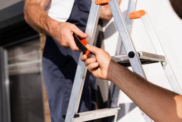 Cropped view of builder giving pliers to colleague on ladder outdoors - Photo, Image