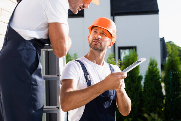 Selective focus of builder in overalls holding digital tablet near colleague on ladder and house  - Photo, Image
