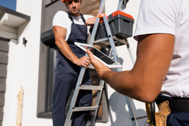 Selectieve focus van handarbeider met digitale tablet naast collega op ladder en gebouw buiten  - Foto, afbeelding