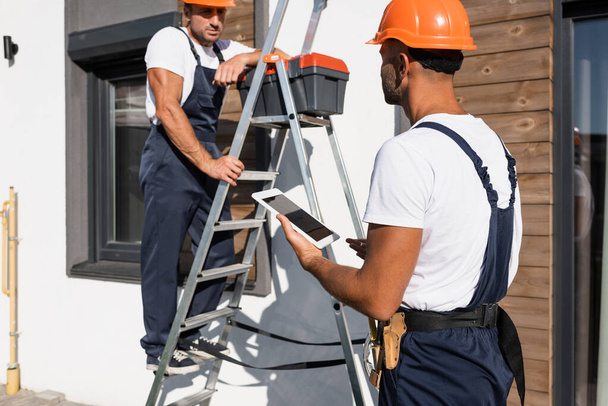 Selective focus of handyman with digital tablet standing beside colleague in uniform on ladder and house  - Photo, Image