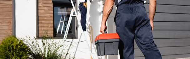 Panoramic crop of builder in uniform holding toolbox near house outdoors  - Photo, Image