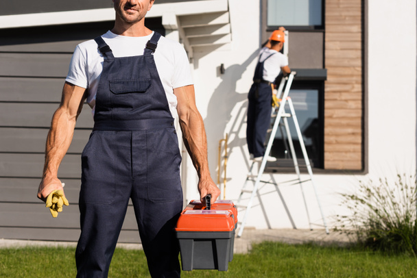 Selective focus of handyman in overalls holding gloves and toolbox on lawn near house  - Photo, Image