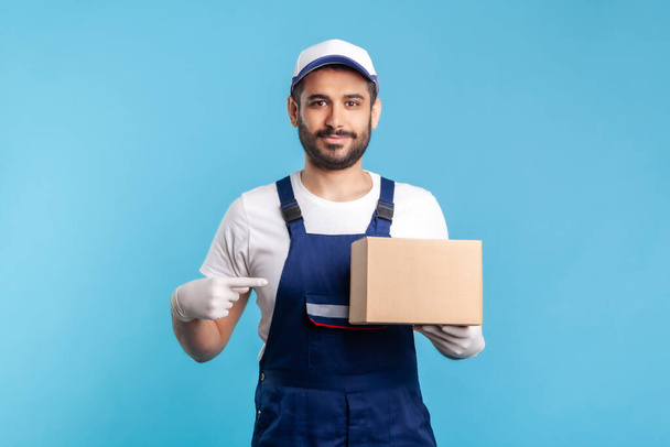 Cargo transportation, delivery service. Friendly worker in uniform pointing at cardboard box and smiling to camera. Courier carrying parcel. Online express shipping, relocation service. isolated - Foto, imagen