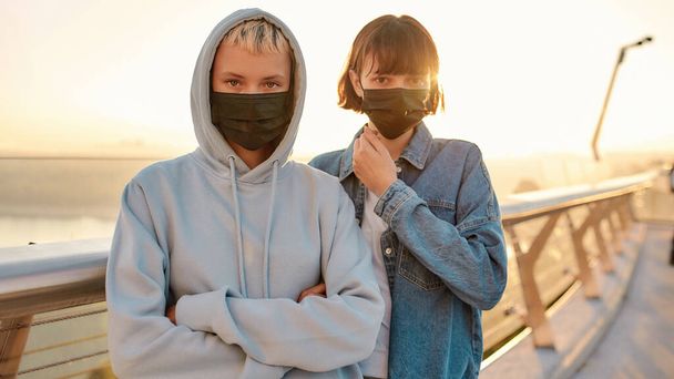 Safety is a choice. Lesbian couple wearing mask, looking at camera, standing on the bridge while admiring the sunrise together - 写真・画像