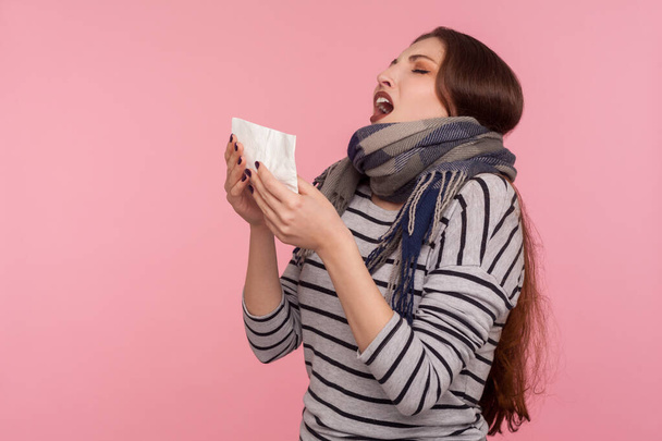 Portrait of unhealthy woman wrapped in warm scarf sneezing in napkin, suffering allergy or flu symptoms, seasonal influenza epidemic, viral infection. indoor studio shot isolated on pink background - Фото, зображення