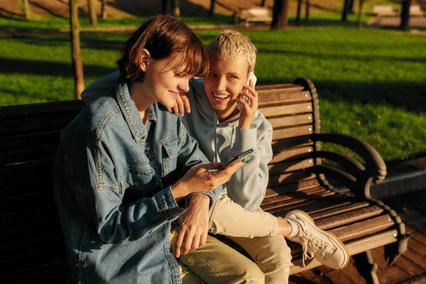 Two joyful girls using smartphone, sitting on the bench in the city park. Lesbian couple holding their phones while spending time together outdoors - Фото, изображение