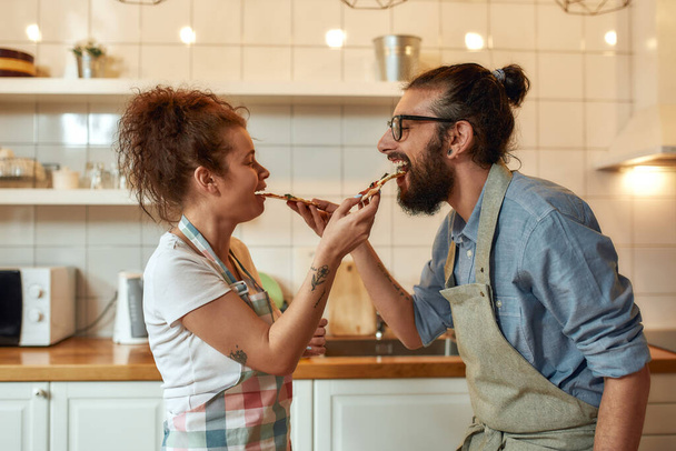Enjoy the taste. Young man and woman in apron feeding freshly baked pizza to each other while standing in the kitchen. Love, relationships concept - Foto, Imagem