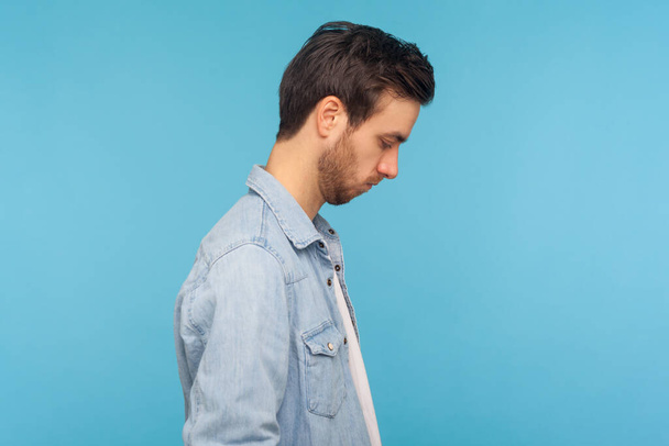 Side view of unhappy man in worker denim shirt looking down, bowing head with sad frustrated expression, feeling depressed, upset about troubles. indoor studio shot isolated on blue background - Photo, image