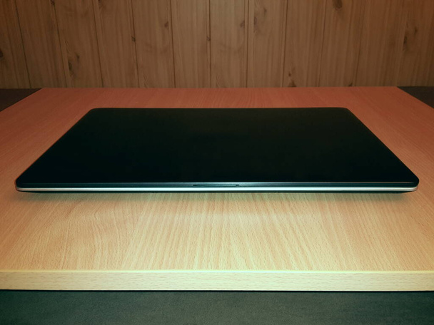 La parte delantera de un ordenador portátil cerrado se encuentra en un escritorio de madera, en el fondo hay una pared de paneles de madera, imagen - Foto, Imagen