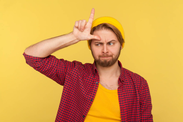 I am so unlucky! Depressed hipster guy in beanie hat and checkered shirt showing L sign, loser gesture on forehead, looking sorrowful frustrated, upset over defeat. indoor studio shot isolated - Фото, зображення