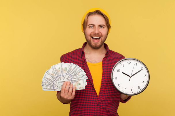 Time to make money! Happy cheerful hipster bearded guy in checkered shirt holding big clock and dollars banknotes, looking at camera smiling satisfied. indoor studio shot isolated on yellow background - Fotoğraf, Görsel
