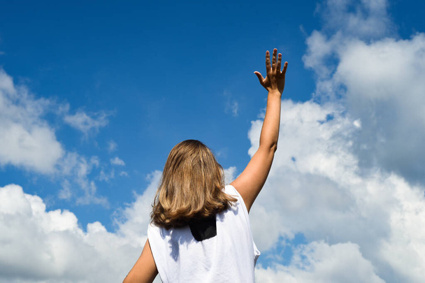 A woman stands with her back to the photographer in a white T-shirt and against a blue sky with clouds. Reaches her hand to the sky.  - Foto, Imagem