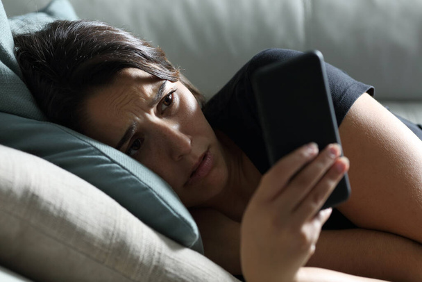 Sad woman checking phone in the dark lying on a sofa in the living room at home - Photo, Image
