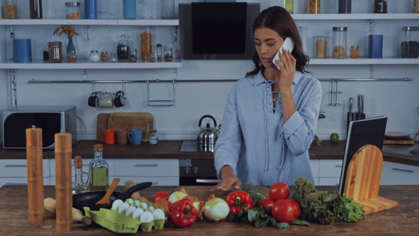 Mujer hablando en smartphone cerca de verduras y huevos en la mesa de la cocina  - Imágenes, Vídeo