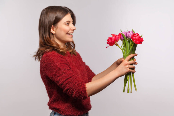 Side view portrait of cheerful happy brunette girl giving flowers bouquet and smiling excitedly. Congratulations on spring holidays, 8 March Women's Day. indoor studio shot isolated on gray background - Фото, изображение
