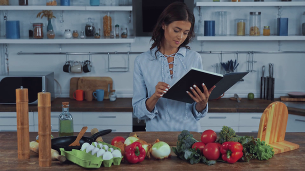 Женщина читает книгу возле свежих овощей на кухонном столе  - Кадры, видео