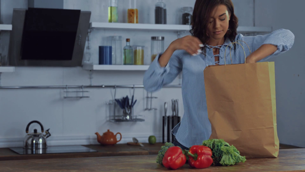 Mujer joven poniendo verduras orgánicas en bolsa de papel en la mesa de la cocina  - Imágenes, Vídeo