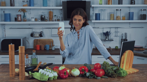 Mutfak masasında taze sebzelerin yanında akıllı telefonuyla selfie çeken bir kadın.  - Video, Çekim