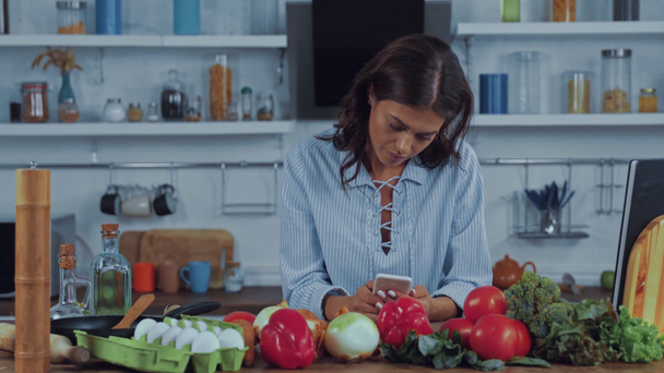 Giovane donna utilizzando smartphone vicino verdure fresche in cucina  - Filmati, video