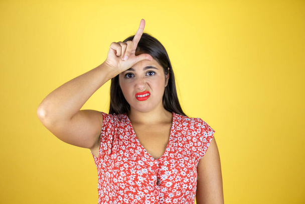 Young beautiful woman over isolated yellow background making fun of people with fingers on forehead doing loser gesture mocking and insulting. - Photo, Image