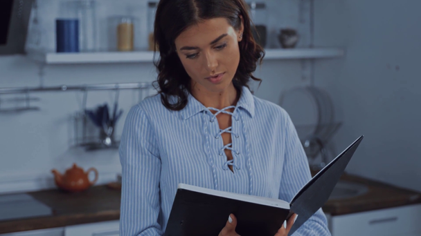 młoda brunetka kobieta czytanie książki w kuchni  - Materiał filmowy, wideo