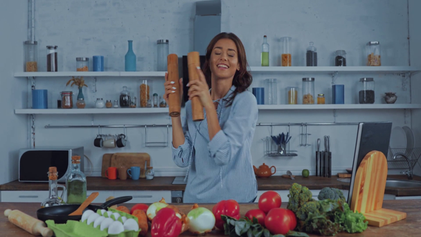 Mujer bailando con pimienta y molinos de sal cerca de verduras en la cocina  - Imágenes, Vídeo