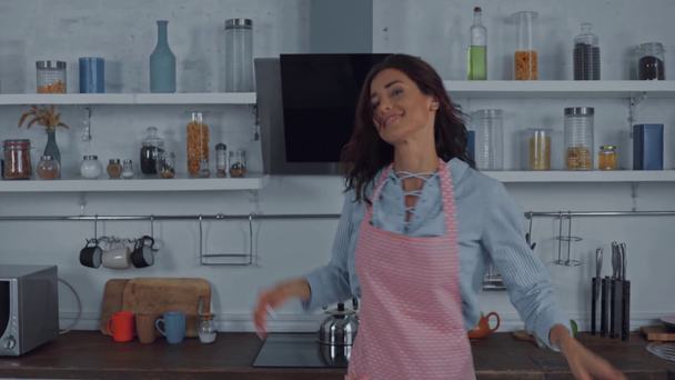 Γυναίκα με ποδιά χορεύει και φυσάει αέρα φιλί στην κουζίνα  - Πλάνα, βίντεο