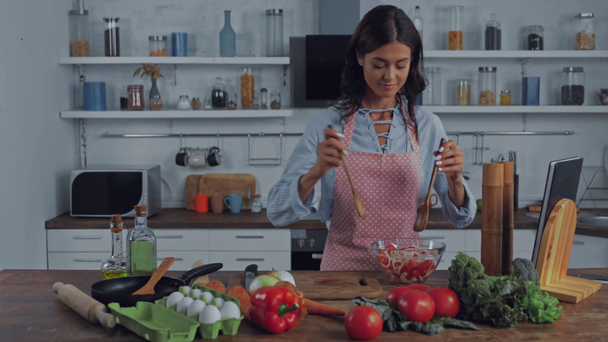 Mujer agregando aceite en ensalada fresca cerca de verduras y huevos en la mesa de la cocina  - Imágenes, Vídeo