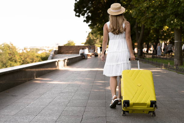 A beautiful tourist girl travels with a yellow suitcase in the park. copy space. - Valokuva, kuva