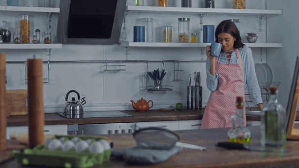 Vrouw in schort drinken koffie in de buurt van ingrediënten in de keuken  - Video