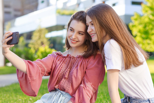Δύο κορίτσια μαθήτριες, το καλοκαίρι σε ένα πάρκο της πόλης, βγάλτε μια selfie σε ένα smartphone, φορώντας καθημερινά ρούχα, ένα διάλειμμα από το σχολείο και το κολέγιο, - Φωτογραφία, εικόνα