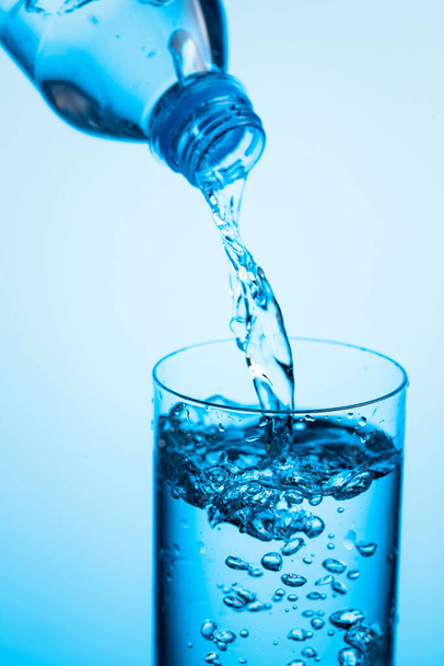 Ρίχνουμε νερό από ένα μπουκάλι σε ποτήρι σε μπλε φόντο. Αντιγραφή χώρου. - Φωτογραφία, εικόνα