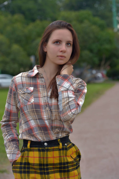 beautiful slender brunette girl in a plaid skirt in the summer in the park - Photo, image