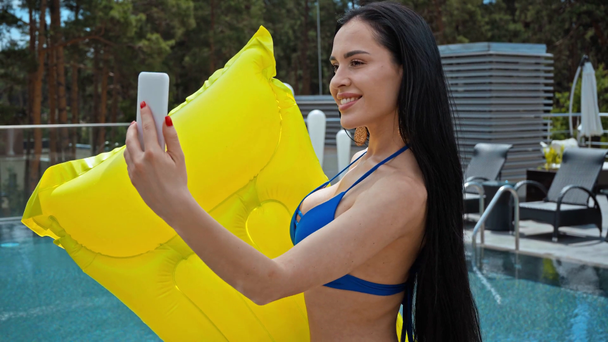 morena mujer en traje de baño tomando selfie con colchón inflable cerca de la piscina  - Metraje, vídeo