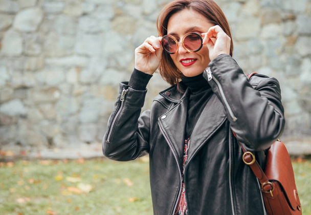 Smiling female dressed boho fashion style in a black leather biker jacket with brown leather flap bag having an autumn park walking. - Φωτογραφία, εικόνα