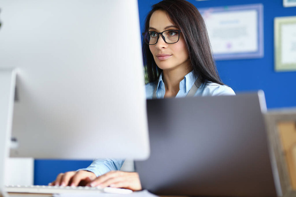 Woman in glasses and blue shirt sit at workplace and look at computer screen. - Foto, afbeelding