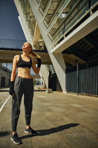 Long shot of young woman who is enjoying sunny rays and closing her eyes after hard training outdoor - Photo, Image