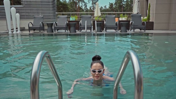 брюнетка в солнцезащитных очках, плавающая в бассейне  - Кадры, видео