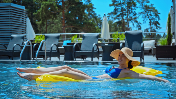 mulher no chapéu de palha acenando a mão e nadando no colchão inflável na piscina  - Filmagem, Vídeo
