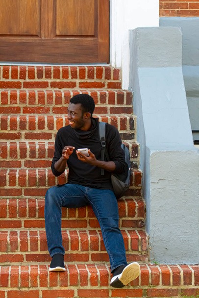 Annapolis, MD 21.8.2020: Ein junger afroamerikanischer Mann mit schwarzem T-Shirt, Jeans, Turnschuhen und Rucksack sitzt auf Ziegeltreppen vor einem Haus und sendet SMS über sein Smartphone.. - Foto, Bild