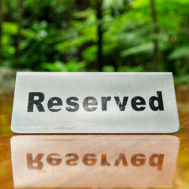 Sinal reservado feito de chapa de aço inoxidável em uma mesa de madeira laminada de um restaurante com árvores e floresta no fundo. - Foto, Imagem