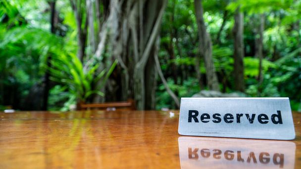 Señal reservada hecha de placa de acero inoxidable sobre una mesa de madera laminada de un restaurante con árboles y bosque en el fondo. - Foto, Imagen