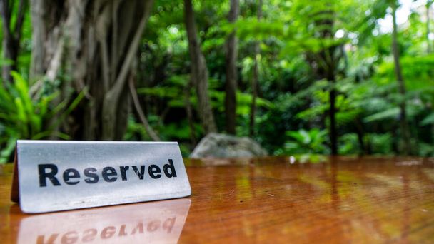 Señal reservada hecha de placa de acero inoxidable sobre una mesa de madera laminada de un restaurante con árboles y bosque en el fondo. - Foto, imagen
