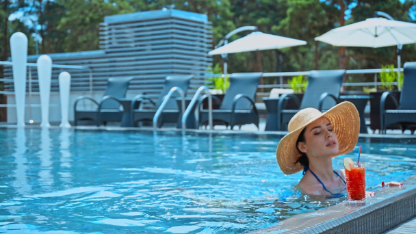 donna in cappello di paglia nuotando in piscina e tenendo il bicchiere con cocktail - Filmati, video