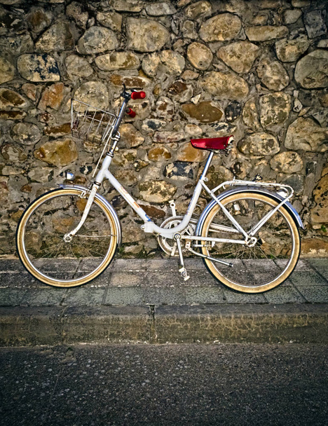 Παλιά λευκό ποδήλατο, με μια ωραία κόκκινη σέλα στηρίζεται σε έναν πέτρινο τοίχο - Φωτογραφία, εικόνα