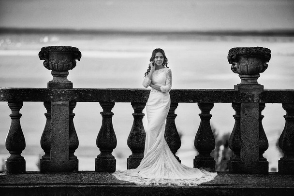 Μια όμορφη νύφη σε ένα πολυτελές φόρεμα στέκεται σε ένα μεγάλο μπαλκόνι. Ασπρόμαυρη φωτογραφία. - Φωτογραφία, εικόνα
