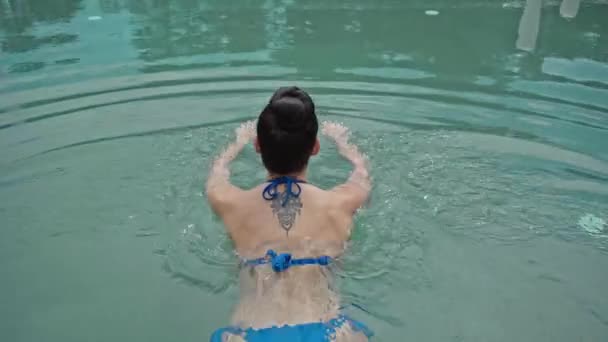 back view of tattooed woman standing with hand on hip and swimming in pool - Filmati, video