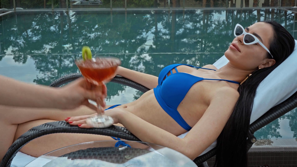 σερβιτόρα που φέρνει κοκτέιλ σε γυναίκα με μαγιό χαλαρώνοντας στην ξαπλώστρα κοντά στην πισίνα - Πλάνα, βίντεο