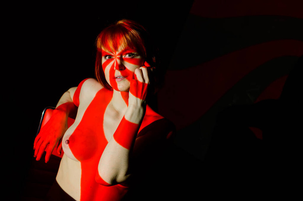 Portrait of a beautiful naked woman in red beams of the projector. Streaks of light and shadow on the skin of a red-haired woman. - Foto, imagen